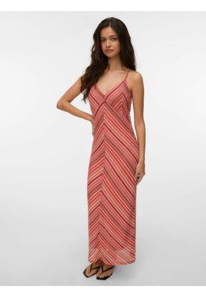 Kvinde med langt mørkt hår iført en ærmeløs maxikjole med røde og pink striber og sorte klipklappere, står med den ene hånd på hoften.