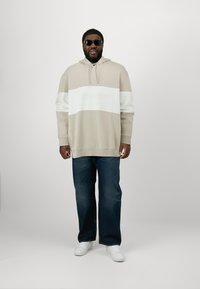 Beige Kapuzenpullover mit weißem horizontalem Streifen, Kordelzugkapuze und Markenlogo. Kombiniert mit dunkelblauen Jeans und weißen Sneakers. Strukturierter Stoff.