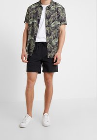 Mies, joka on pukeutunut mustiin shortseihin, valkoisiin tennareihin, valkoiseen t-paitaan sekä auki olevaan lyhythihaiseen nappipaitaan vihreällä palmulehtikuvioinnilla.