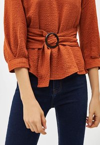 Kobberfarvet bluse med struktur og 3/4-ærmer, der er bundet i taljen med et bredt bælte og en rund sort spænde, parret med mørkeblå jeans.