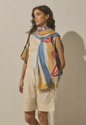 Woman wearing beige sleeveless top and shorts, with a multicolored scarf and woven shoulder bag, standing against a plain wall.