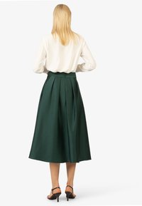 Groene geplooide midirok met een gladde textuur en aansluitende tailleband, gecombineerd met een witte blouse en zwarte hoge hakken sandalen.
