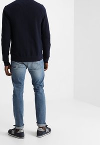 Marinblå tröja, ljusblå jeans och grå New Balance-sneakers. Tröjan har rundad halsringning; jeansen är raka med uppvikta ben.