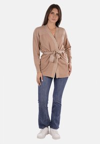 Cardigan beige lavorato a maglia con texture a coste, apertura frontale e cintura da annodare. Indossato sopra jeans a zampa d'elefante blu e sneakers bianche.