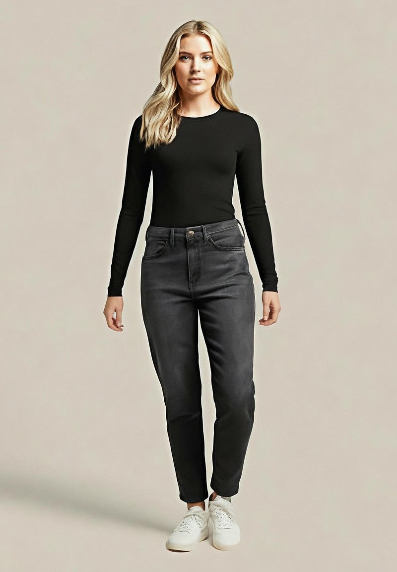 Blondynka stoi w czarnym długim rękawie topie, szarych spodni z wysokim stanem i białych sneakersach na jednolitym beżowym tle.