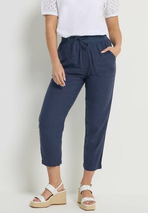 Kvinde iført marineblå bukser med snor, hvid top med blonder på ærmerne og hvide kilehælssandaler, stående foran en ensfarvet baggrund.