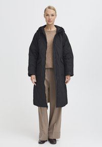 Sort quiltet lang jakke med hætte, der har lynlås foran, to sidelommer og et struktureret mønster. Båret over en lys sweater og bukser.