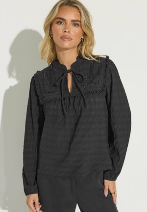 Blondynka nosi czarną, teksturowaną bluzkę z długim rękawem, wykończoną falbankami i koronkowymi zdobieniami, stojąc na tle jasnego jednolitego tła.