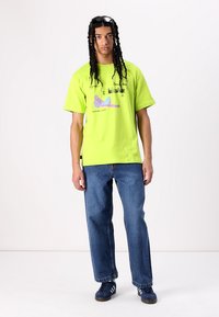 Neongrønn bomull t-skjorte med grafisk trykk, sammen med løse blå denimjeans og marineblå sneakers med tre hvite striper.
