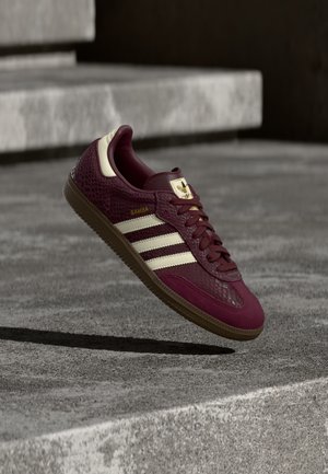 Burgundy sneaker with textured leather, three white stripes, and gold accents. Rubber sole, classic Samba design, and breathable mesh details.