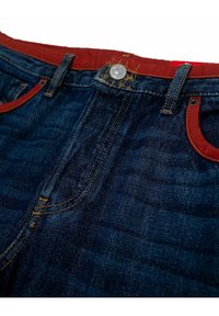 Farkut tummansinisellä pesulla, joissa on punaiset yksityiskohdat taskuissa ja vyötärönauhassa, näkyvät ompelusaumat ja metallinen nappiyksityiskohta.