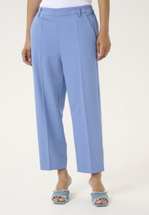 Vrouw draagt lichtblauwe hoge taille broek met voorplooien en zijzakken, gecombineerd met open teen denim slip-on hakken.