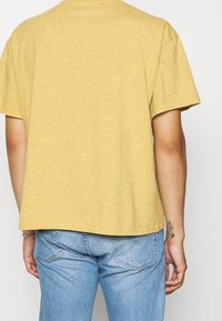 Gul bomullströja med avslappnad passform, korta ärmar och rundad halsringning, visad bakifrån. Har en liten tatuering på handleden.