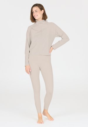 Beige Sportanzug mit hochgeschlossenem Reißverschluss und langen Ärmeln sowie eng anliegenden Leggings, gefertigt aus glattem, dehnbarem Material mit geripptem Textur.