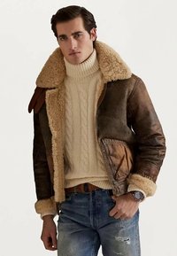 Man med brun shearling läderjacka, krämfärgad kabelstickad polotröja, blå slitna jeans och klocka, stående mot en neutral bakgrund.