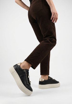 Persoon draagt donkerbruine ribfluwelen broek en zwarte plateau sneakers met witte zolen en gouden hielaccenten, heft één voet op.