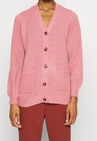 Cardigan in maglia rosa con scollo a V, texture a coste, chiusura con sei bottoni e due tasche frontali. Presenta maniche a palloncino e bottoni lucidi.