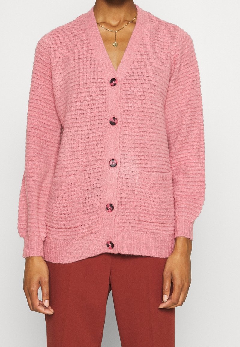 Cardigan in maglia rosa con scollo a V, texture a coste, chiusura con sei bottoni e due tasche frontali. Presenta maniche a palloncino e bottoni lucidi.