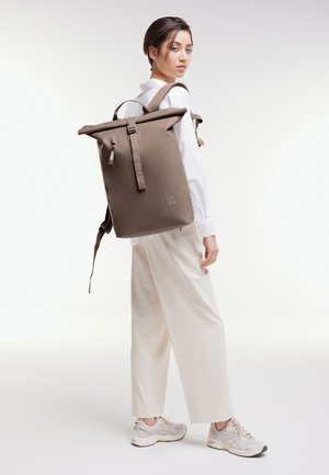 Vrouw met een wit shirt, brede beige broek en sneakers, draagt een grote bruine roll-top rugzak op haar rug, zijwaarts kijkend.