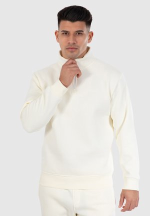 Cremefarvet pullover i blødt stof med høj krave, halvlukning med lynlås og ribbede manchetter og kant. Har diskret branding.