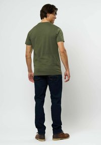 Vyras atsisukęs nugarą, dėvintis alyvuogių žalią trumpomis rankovėmis marškinėlį, tamsiai mėlynas džinsus ir rudos odinės batai prieš paprastą šviesią foną.