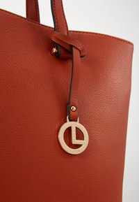 Sac fourre-tout en cuir rouge texturé avec un accent noir et un charm logo circulaire doré attaché à la sangle. Poignées lisses et finition polie.
