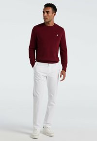 Bordeaux gebreide trui met ronde hals, gecombineerd met witte op maat gemaakte broek. Draagt witte sneakers en staat tegen een effen achtergrond.