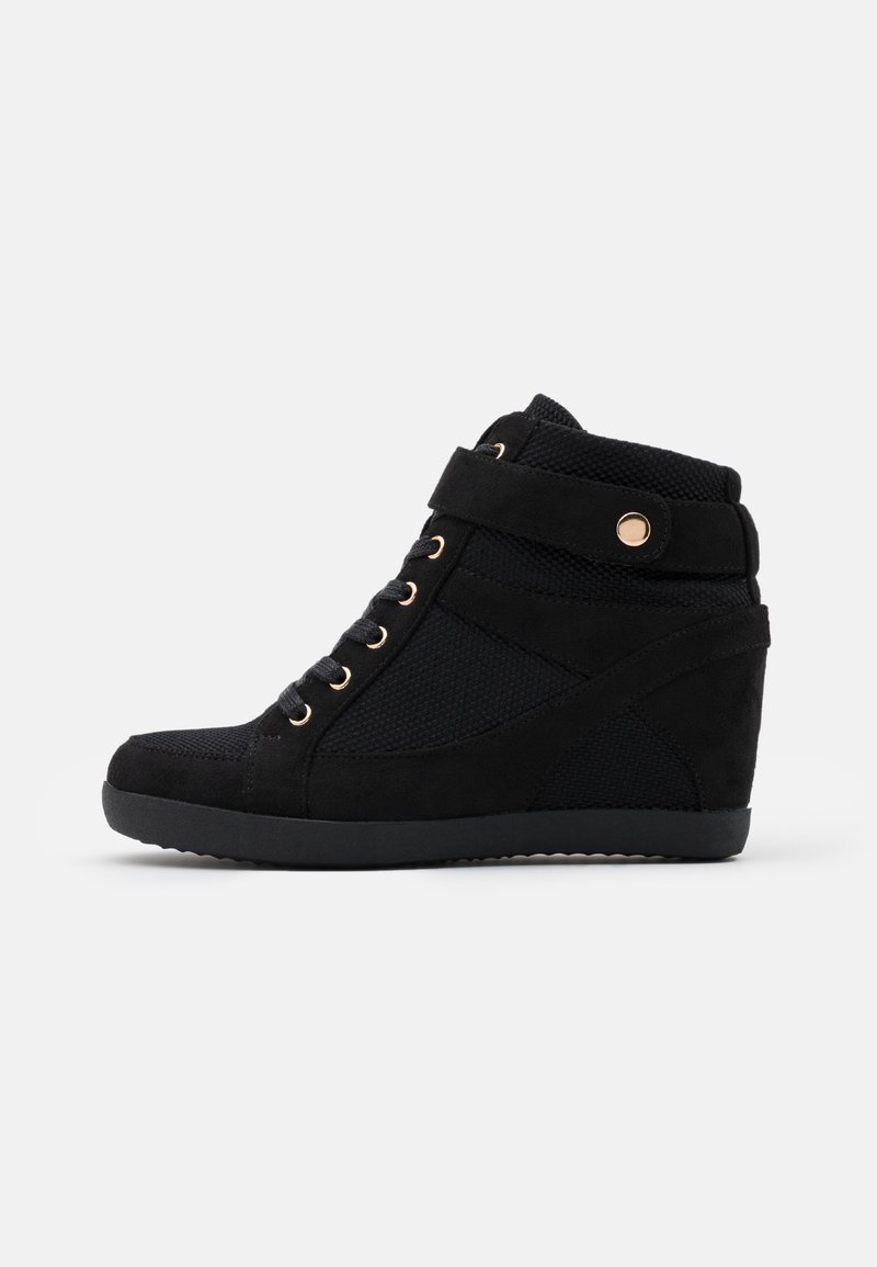 Black wedge sneakers with suede and mesh material, featuring gold eyelets, a strap across the ankle, and a textured sole.
