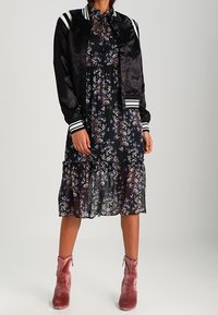 Musta satiininen bomber-takki valkoisilla raidoilla kukkakuosisen mustan midi-mekon päällä. Vaaleanpunaiset samettiset nilkkurit täydentävät asukokonaisuuden.