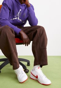 Zapatillas Nike blancas con acentos rojos y perforaciones, combinadas con pantalones marrones y una sudadera morada con capucha que presenta un logo gráfico.