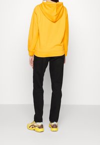 Sudadera amarilla con capucha, pantalones negros y zapatillas amarillas y crema. La sudadera tiene un bolsillo tipo canguro y un corte relajado.