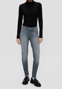 Vaqueros ajustados de denim azul desgastado, con cintura de altura media, bolsillos delanteros y cierre con botones. Combinados con un suéter de cuello alto negro.