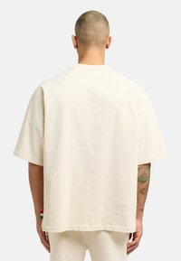 Beige overdimensjonert t-skjorte med rund hals og korte ermer, utstyrt med et preget logo på baksiden og en liten farget etikett på siden.