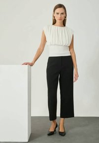 White, textured top with pleats and a fitted waist, paired with black cropped trousers and black heels, set against a neutral background.