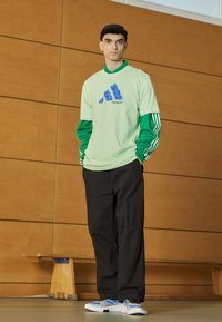 Camicia verde a maniche lunghe sotto una camicia verde chiaro a maniche corte con il logo Adidas blu, pantaloni neri e scarpe bianche con dettagli blu.
