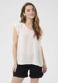 Blouse blanche légère avec un décolleté en V et des manches à volants. Texture douce, légèrement transparente avec des rayures verticales. Portée avec un short noir retroussé.