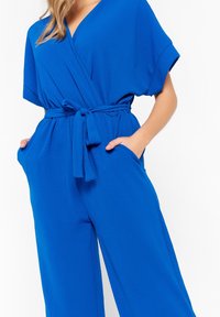 Niebieski kombinezon z miękkiego materiału, z szerokimi krótkimi rękawami, w talii z paskiem oraz bocznymi kieszeniami. Gładka tekstura i luźny krój.