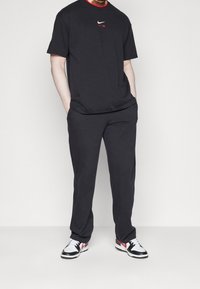 Man wearing black Nike Air t-shirt, black pants, and white sneakers with black and red details, hands in pockets, standing against white background.