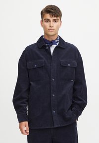 Navy corduroy shirt jacket with two front pockets, button closure, and a spread collar. Textured fabric with vertical ribbing, casual fit.