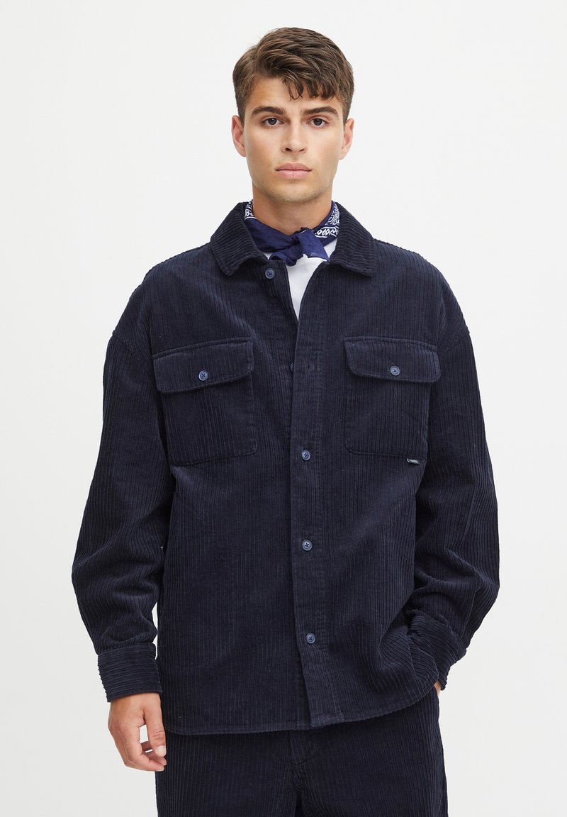 Navy corduroy shirt jacket with two front pockets, button closure, and a spread collar. Textured fabric with vertical ribbing, casual fit.