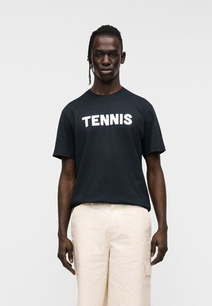 Schwarzes Baumwoll-T-Shirt mit weißem "TENNIS" Schriftzug, kurzen Ärmeln, lockerem Schnitt. Dazu trägt man hellbeige Hosen mit einfarbiger Textur und geradem Schnitt.