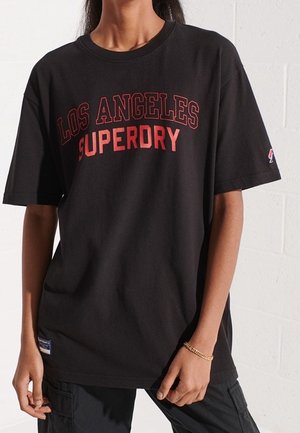 Osoba nosząca czarną oversize'ową koszulkę z czerwonym napisem "LOS ANGELES SUPERDRY", w połączeniu z czarnymi spodniami i złotą bransoletką.