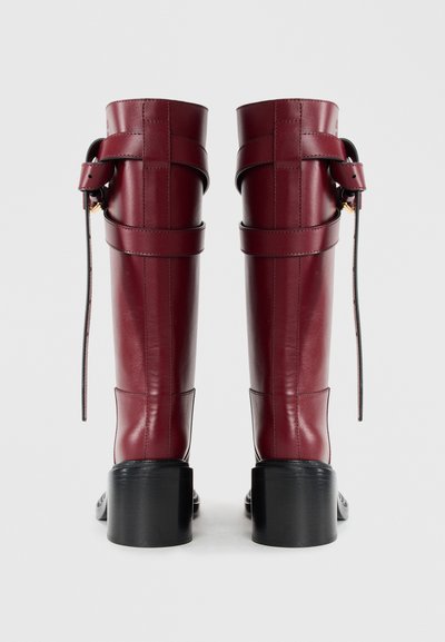 Burgundy leather knee-high boots with a wide cuff and adjustable strap. Textured finish and block heel enhance structural design.