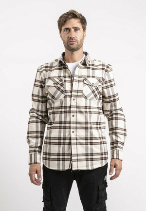 Homme aux cheveux bruns courts portant une chemise à carreaux beige boutonnée par-dessus un t-shirt blanc et un pantalon cargo noir, debout devant un fond uni.