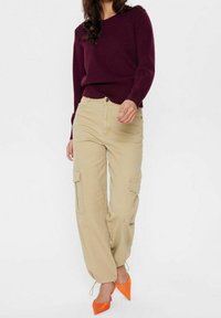 Kobieta w ciemnofioletowym swetrze z dzianiny, beżowych spodniach cargo z kieszeniami oraz jasnopomarańczowych szpilkach z ostrym noskiem, stojąca na tle jednolitym.