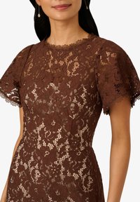 Robe en dentelle marron avec des manches courtes et volantées, un col festonné, des motifs floraux et une silhouette ajustée avec des détails transparents.
