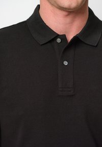 Svart polo-tröja gjord av texturerat tyg med en treknapps krage och en klassisk krage. Funktioner med dämpade grå knappar.