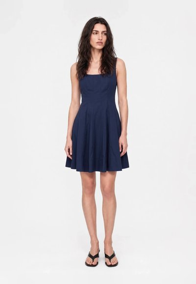 Femme aux longs cheveux foncés portant une robe sans manches bleu marine longueur genou et des sandales noires à talons, debout sur un fond blanc uni.