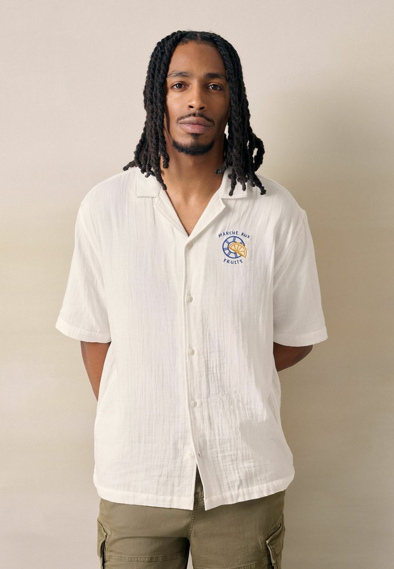 Homme aux dreadlocks épaules, portant une chemise blanche boutonnée avec le logo "Marché Aux Fruits" et un pantalon cargo olive, debout contre un mur beige.