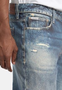 Svijetloplave traperice s izblijedjelim završetkom, s istrošenim detaljima, pet džepova i kožnom oznakom s natpisom "Guess Jeans."
